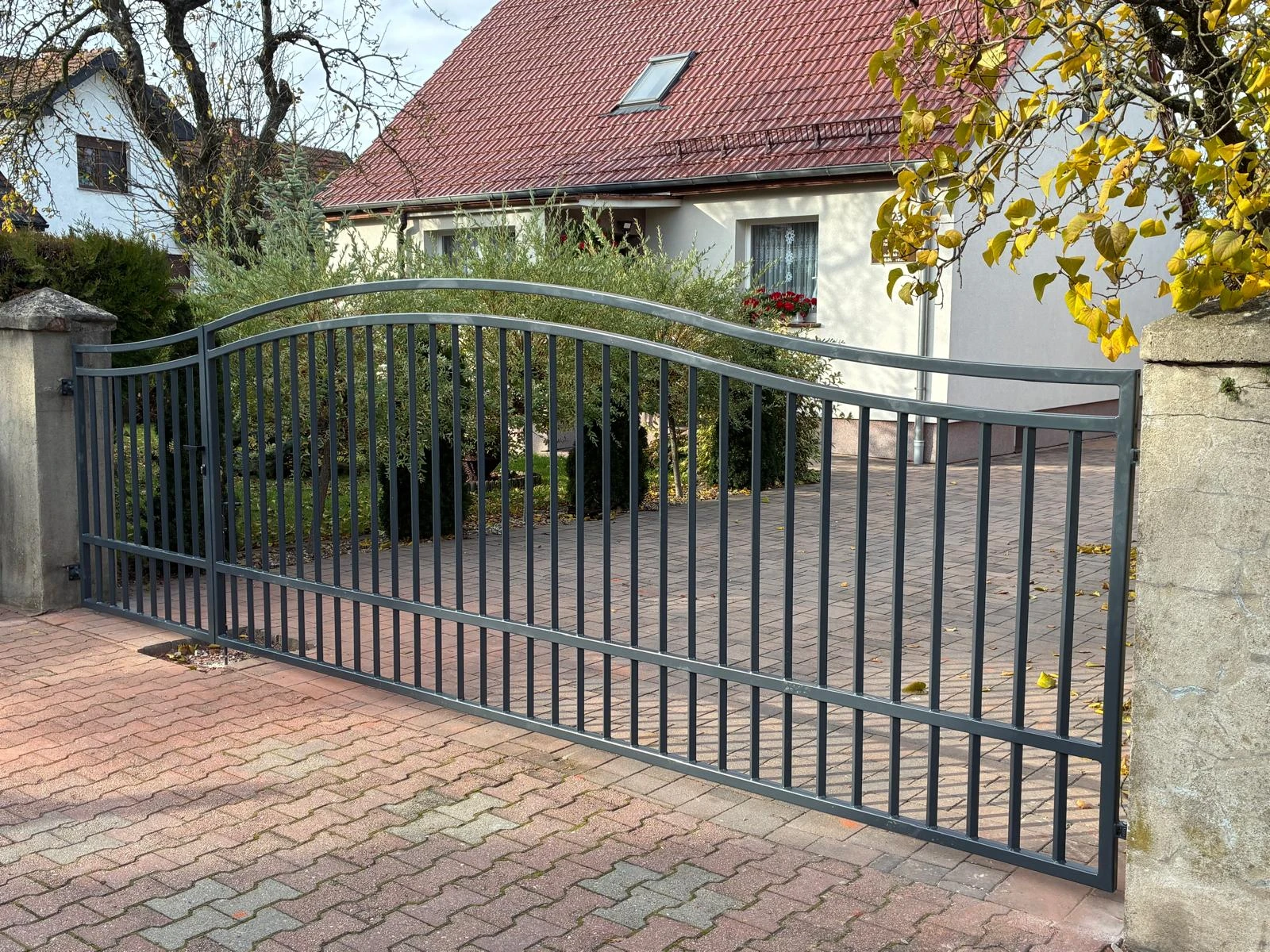 Metalowa brama wjazdowa z pionowymi prętami i łukowym górnym profilem na podjeździe przed domem