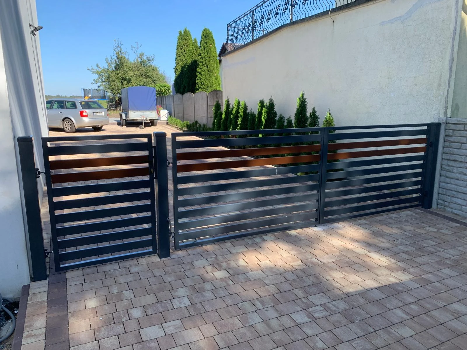 Nowoczesna brama wjazdowa i furtka z metalowymi poziomymi przęsłami na tle podjazdu z kostki brukowej